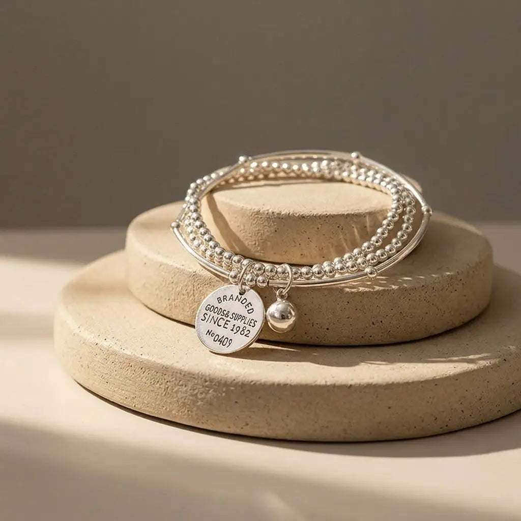 Three-layer silver bead stretch bracelet set with engraved coin charm and silver ball charm, displayed on stacked round sandstone disc props with warm directional sunlight and shadow, warm taupe background

