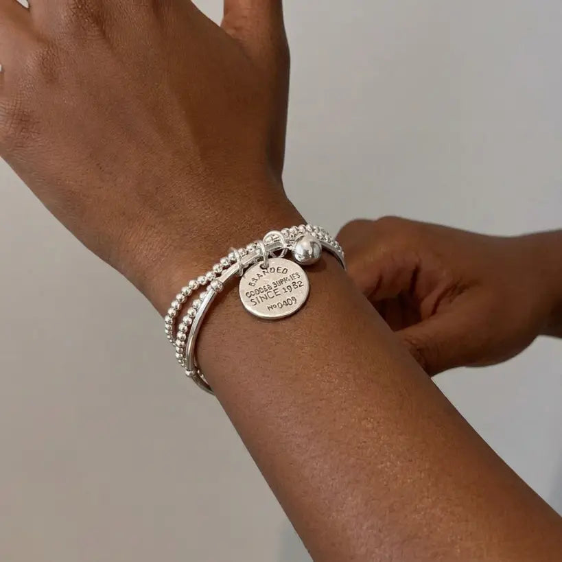 Close-up of deep skin tone woman's wrist wearing three-layer silver bead stretch bracelet set, other hand fingers gently touching bracelet with engraved coin charm and silver ball charm visible, light grey background

