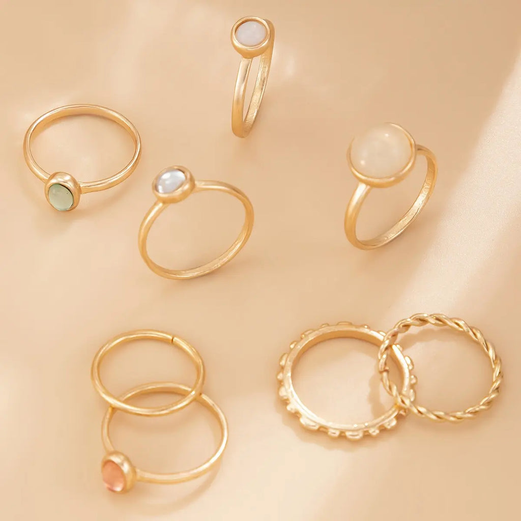 7-piece boho gold-tone alloy stacking ring set scattered on warm beige matte fabric surface — green natural stone ring, white mother-of-pearl ring, peach cat's eye stone ring, plain thin band rings, round bead texture band and twisted rope band rings — warm directional natural light from upper right, AI-generated image

