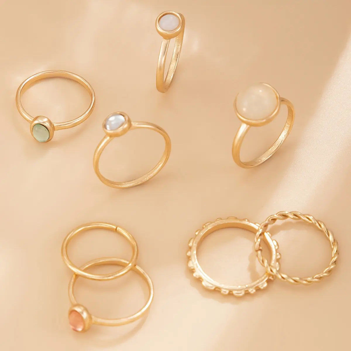 7-piece boho gold-tone alloy stacking ring set scattered on warm beige matte fabric surface — green natural stone ring, white mother-of-pearl ring, peach cat's eye stone ring, plain thin band rings, round bead texture band and twisted rope band rings — warm directional natural light from upper right, AI-generated image

