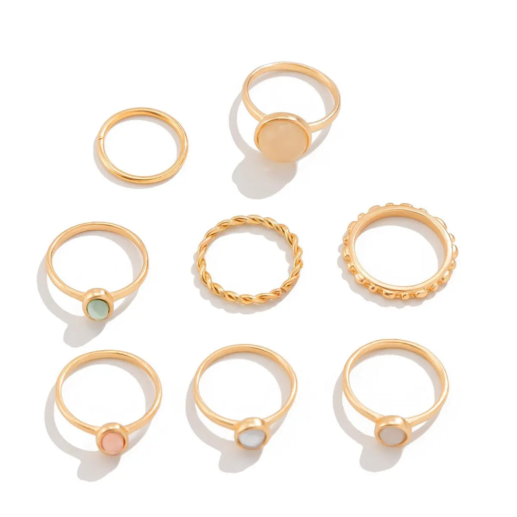 7-piece boho gold-tone alloy stacking ring set displayed on white background: plain thin band ring, large round peach cat's eye stone ring, round green natural stone ring, twisted rope band ring, round bead texture band ring, round pink natural stone ring, and round white mother-of-pearl ring; white background with soft drop shadow

