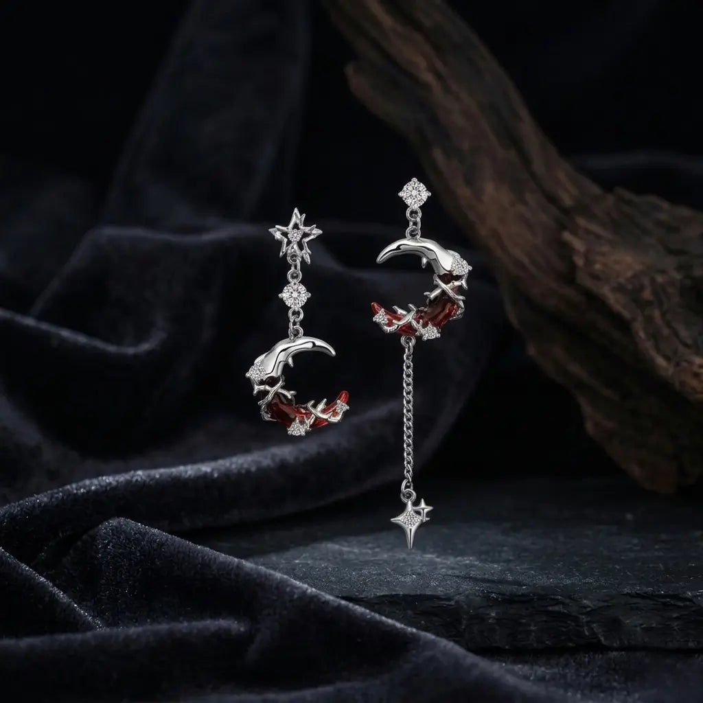 Asymmetric silver-tone moon and star drop earrings displayed on dark black velvet fabric — left short crescent moon earring with red and white zircon thorn detail; right long crescent moon earring with silver chain and four-pointed spike star drop — dark brown driftwood prop in background, soft ambient light, gothic atmospheric display


