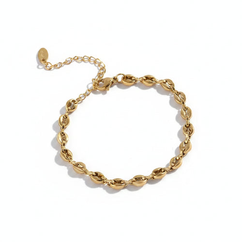 18K gold plated coffee bean chain bracelet with micro-pave zirconia center links, lobster clasp and oval logo tag extender, draped over stacked round perforated sandstone disc props with palm leaf shadow, warm taupe background

