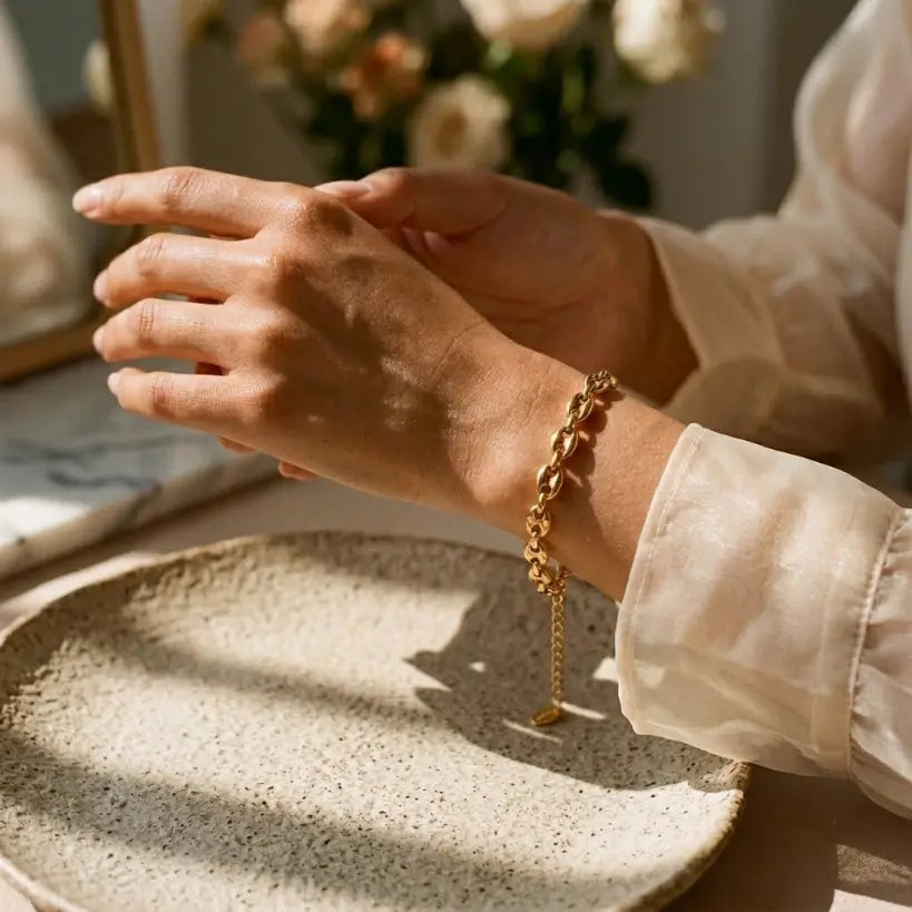 Woman displaying 18K gold plated coffee bean chain bracelet draped across open palm, extender chain and oval logo tag pendant visible, styled with cream organza ruffle sleeve, gold mirror frame and cream rose bouquet in background

