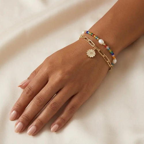 Woman with medium skin tone wearing double bracelet set — multicolor seed bead and pearl strand layered with gold paperclip chain and white daisy enamel charm — on left wrist, hand resting on cream satin fabric in warm natural light


