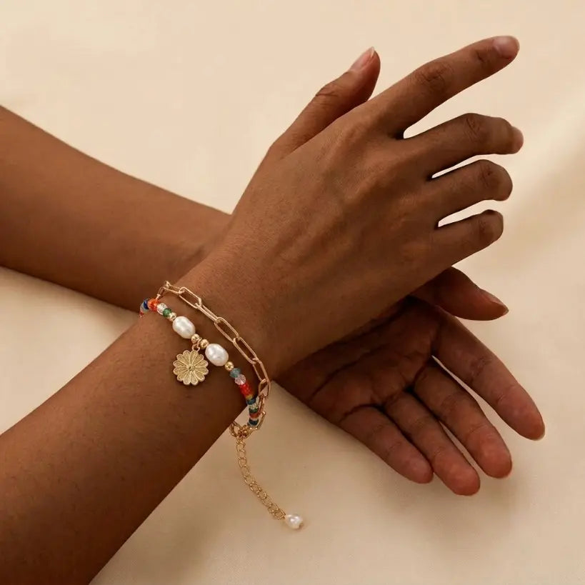Woman with deep skin tone wearing double bracelet set — multicolor seed bead and pearl strand layered with gold paperclip chain and white daisy enamel charm pendant — on left wrist, hands crossed, warm beige background

