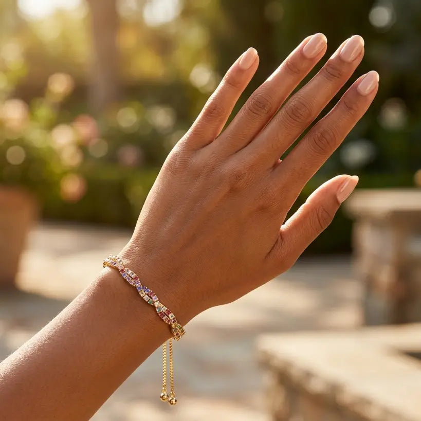 Close-up of hand wearing gold plated multicolor zirconia tennis bracelet with drawstring closure on right wrist, palm facing outward against warm golden sunlight with blurred garden and stone path background

