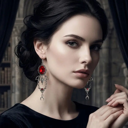 Woman with light skin tone and black updo wearing gothic silver-tone crimson bat and moon drop earrings — deep red oval glass gemstone in silver filigree frame, spread-wing bat pendant, dual chain swags with red crystal connector and silver crescent moon drop — smoky eye makeup, finger touching chin, black round-neck top, dark gothic library with black drape background, AI-generated model image

