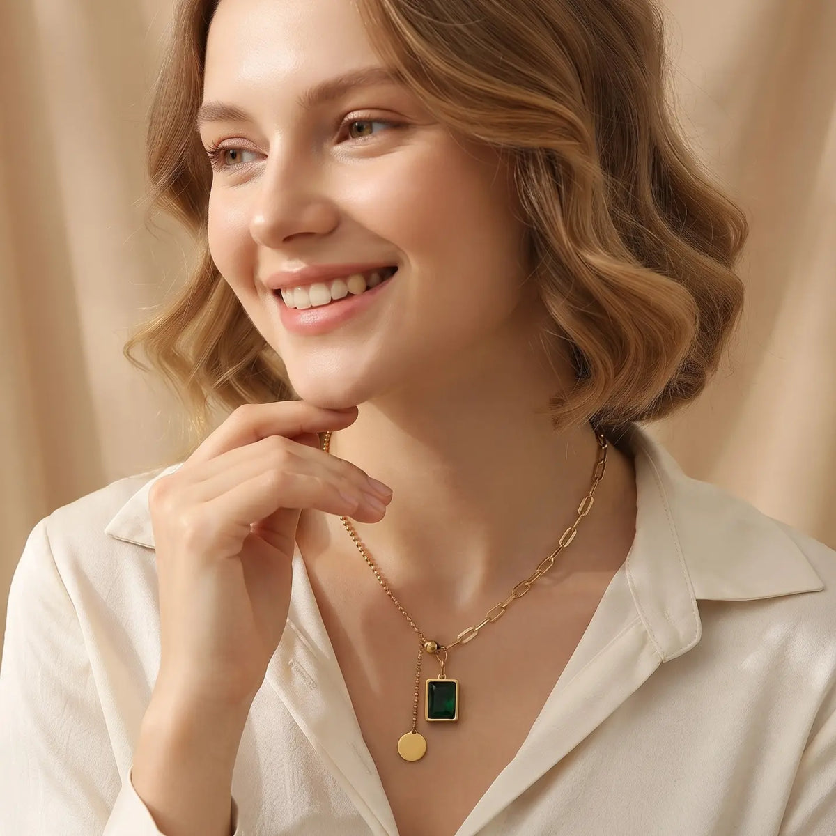 Smiling woman with light skin tone wearing gold mixed chain necklace with emerald green square crystal pendant and round coin charm, dressed in cream collared shirt, warm champagne silk drape background

