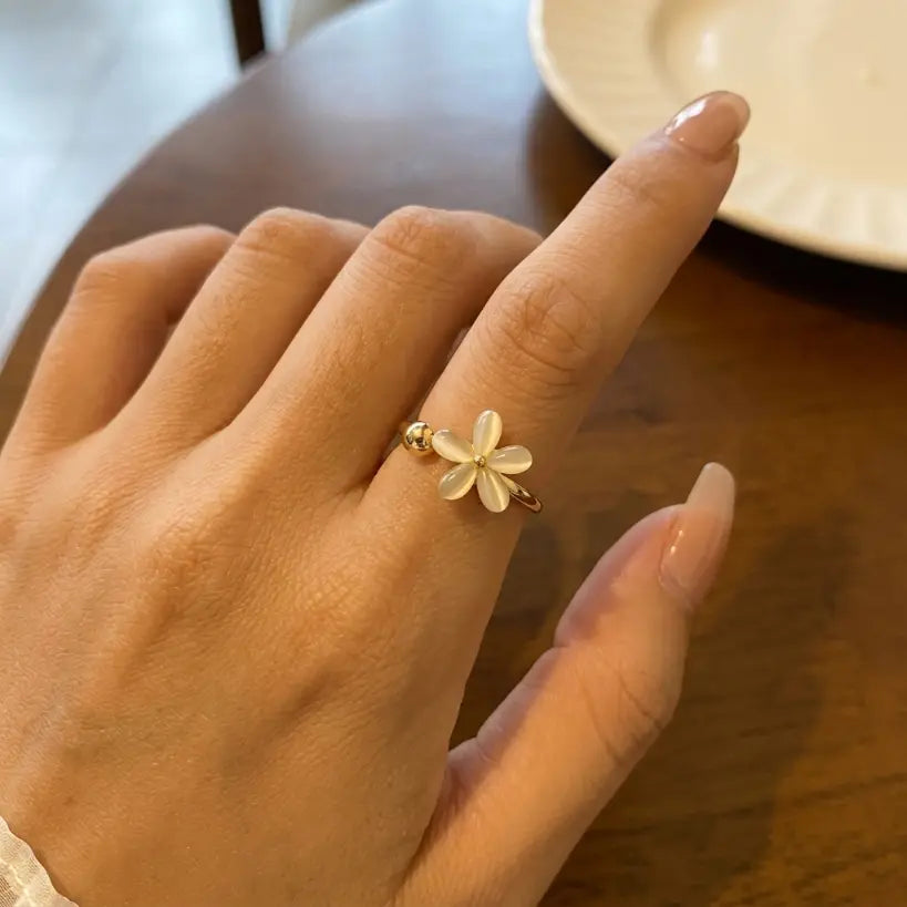 Woman wearing gold plated copper adjustable open band ring with white cat eye five-petal flower and gold bead on ring finger, styled with white lace sleeve, dark wood table background
