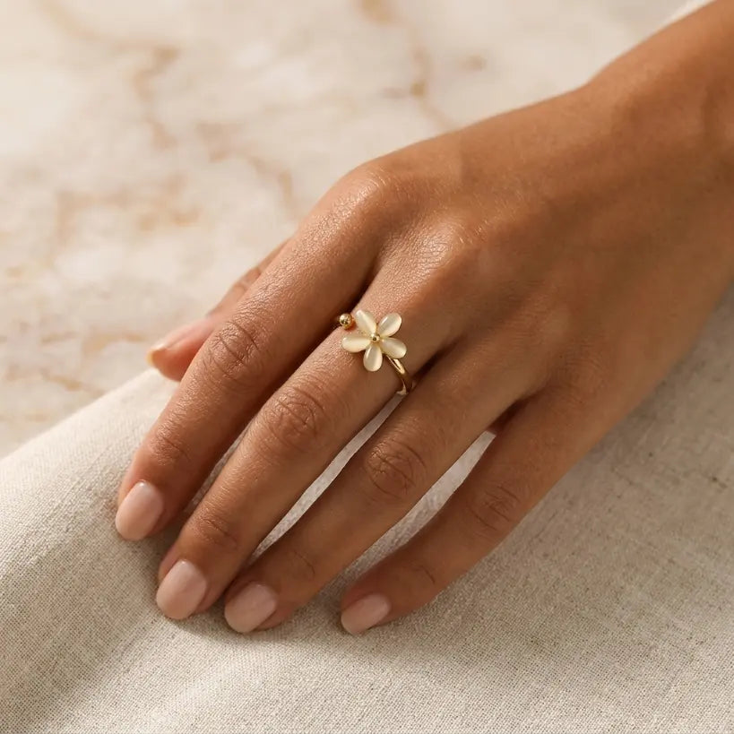 Woman wearing gold plated copper adjustable open band ring with white cat eye five-petal flower and gold bead on middle finger, resting hand on white linen with marble background in soft natural light