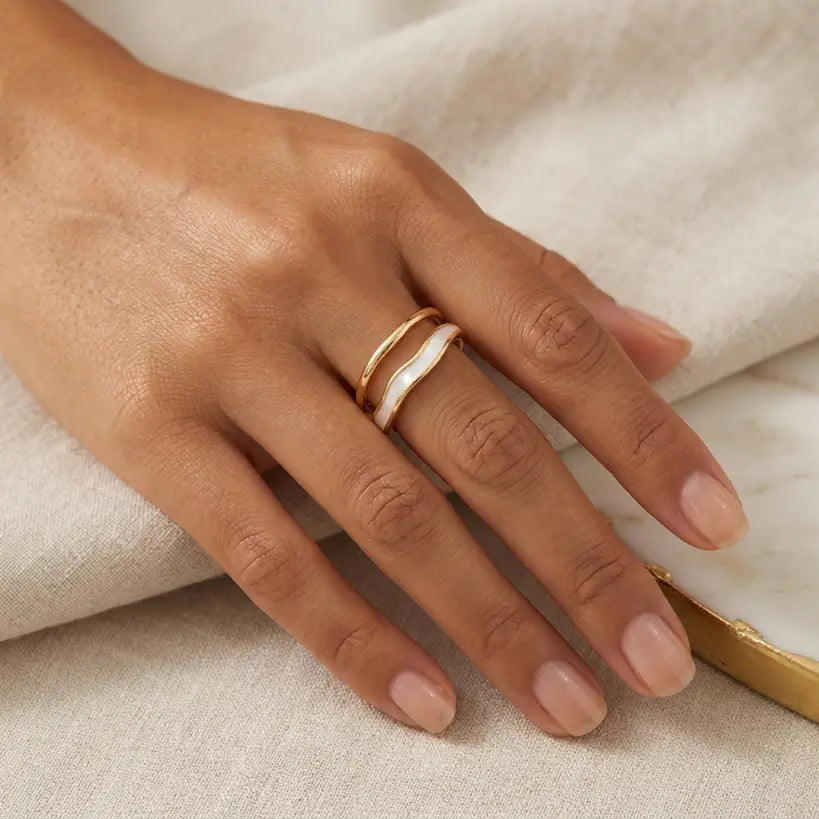 Woman wearing gold double-layer irregular wave open band ring with white enamel inlay on middle finger, hand resting on folded cream linen fabric with gold metal accent in corner, soft natural light

