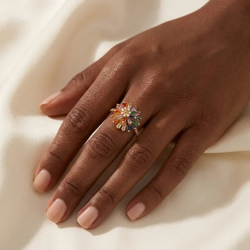 Woman with deep skin tone wearing gold alloy adjustable open band ring with rainbow multicolor baguette zirconia sunflower and pearl center on middle finger, hand resting on cream satin fabric

