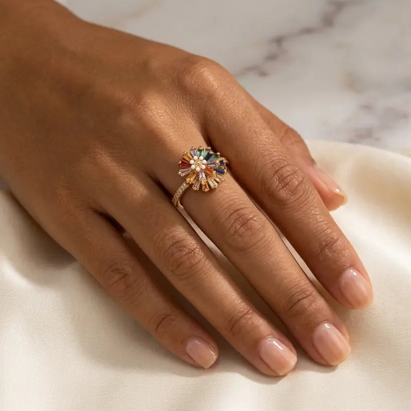 Woman wearing gold alloy adjustable open band ring with rainbow multicolor baguette zirconia sunflower and pearl center on middle finger, hand resting on white satin fabric with marble background

