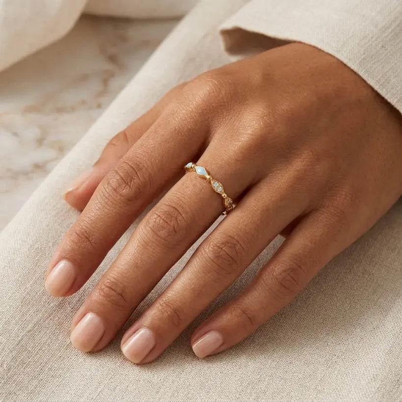 Woman wearing gold alloy adjustable open band ring with light blue diamond-shaped enamel and zirconia accents on middle finger, hand resting on cream linen fabric with marble and white jacket sleeve background

