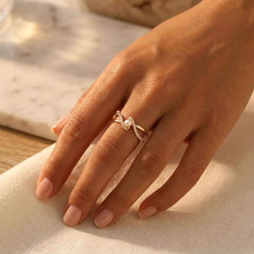 Woman wearing gold alloy adjustable open band ring with double micro-pave zirconia crossover bands and freshwater pearl center on middle finger, resting on white linen with marble background in warm natural light