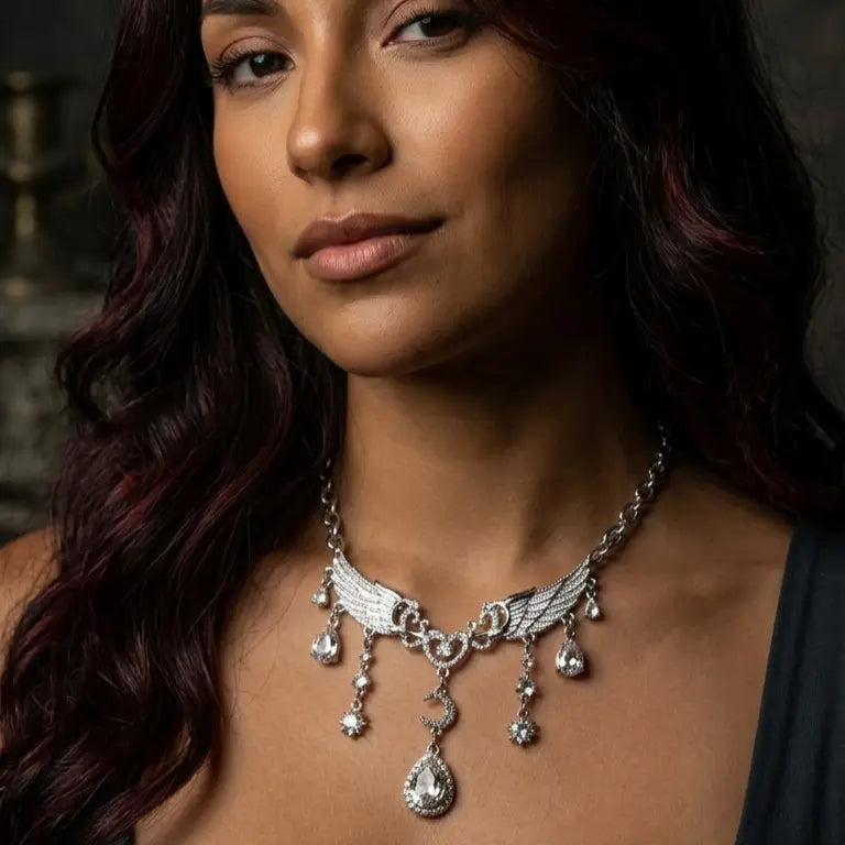 Woman with medium-deep skin tone and long dark auburn wavy hair wearing gothic silver-tone angel wing heart moon rhinestone drop necklace, gaze directed upward, dark grey V-neck top, dark gothic interior with candelabra background, AI-generated model image

