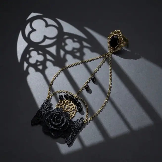 Gothic black lace rose onyx slave bracelet ring set with 3D black fabric rose, antique bronze diamond mesh base, black bead drops and connected filigree black onyx ring, displayed on dark blue-grey concrete surface with dramatic gothic quatrefoil rose window and lancet arch shadow projection, strong directional spotlight, AI-generated image

