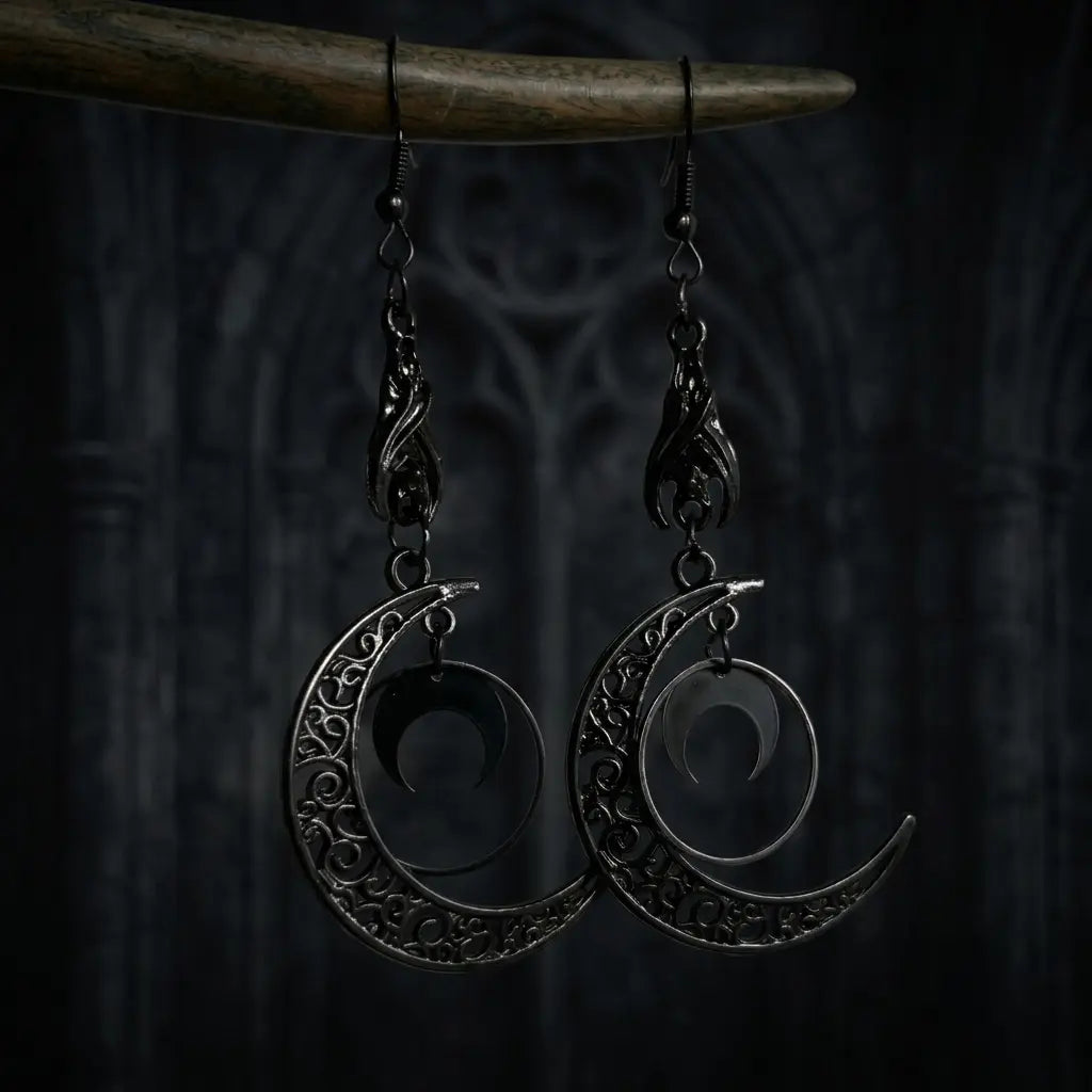 Pair of gothic gunmetal black filigree crescent moon drop earrings hanging from dark brown driftwood branch — black flame claw connector, large hollow scrollwork crescent moon with nested solid crescent inside — dark blue-grey gothic wooden architectural background with lancet arch carved detail, soft ambient light


