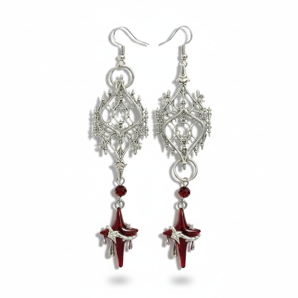 Pair of gothic silver-tone copper alloy filigree drop earrings: silver fish hook, round connector ring, large silver gothic diamond-shaped filigree pendant with lancet arch thorn candelabra cutout detail, silver ring connector, deep red faceted crystal bead, and deep red glass four-pointed star pendant in silver claw setting with three blood drop drips at bottom; isolated on white background with soft drop shadow

