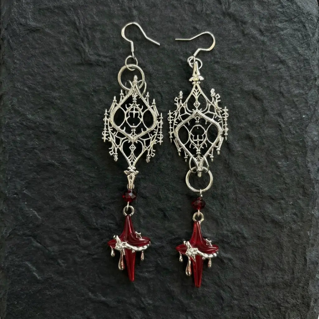 Pair of gothic silver-tone filigree drop earrings displayed flat on dark black rough stone slab — large diamond-shaped gothic lancet arch filigree pendant, deep red faceted crystal bead and deep red glass four-pointed blood star drip drop — soft ambient light, dark atmospheric display

