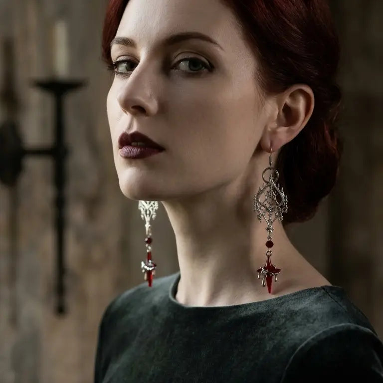 Woman with light skin tone and deep red updo wearing gothic silver-tone filigree drop earrings with large diamond-shaped gothic filigree pendant, deep red crystal bead and blood star drip drop on both ears, deep burgundy lips, dark grey-green round-neck top, dark gothic stone wall with iron wall sconce candelabra background, AI-generated model image

