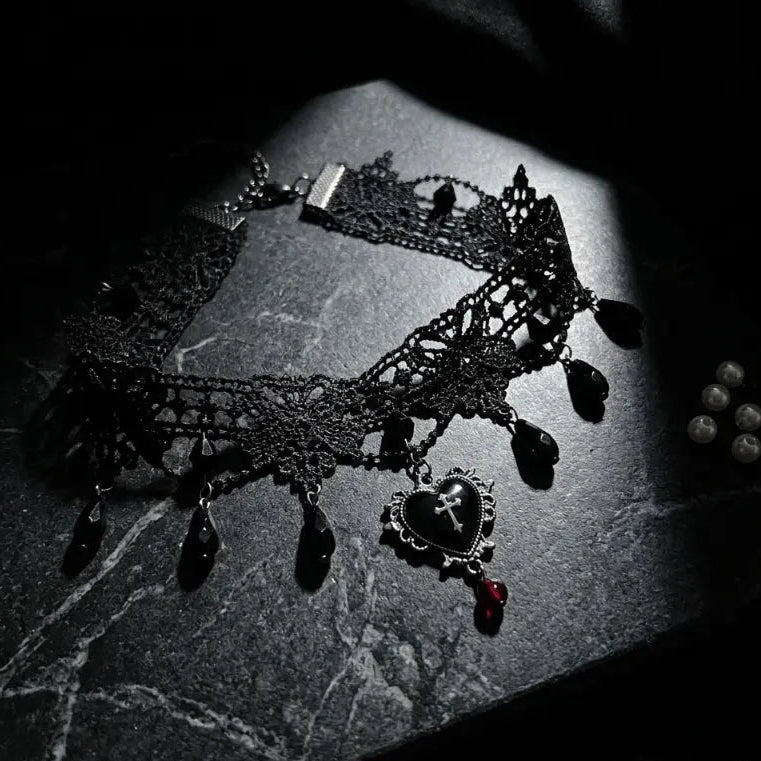 Gothic black lace filigree choker necklace with black teardrop bead drops, silver filigree heart cross pendant and red crystal teardrop drop, displayed on dark grey marble surface with scattered white pearl props, dramatic spotlight, AI-generated image