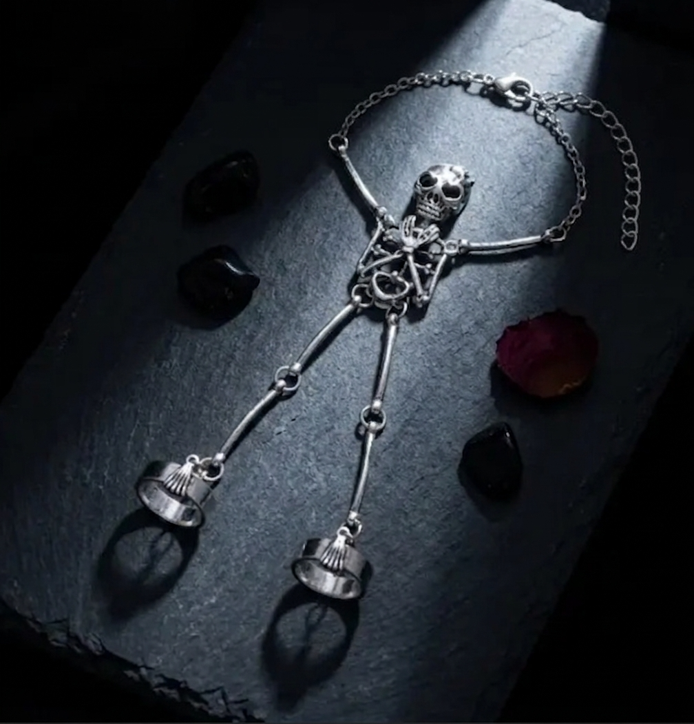 Gothic silver-tone full-body skeleton hand chain bracelet double ring set, displayed on dark blue-grey stone slab with scattered black obsidian stones and a single deep red rose petal, dramatic single spotlight, black background
