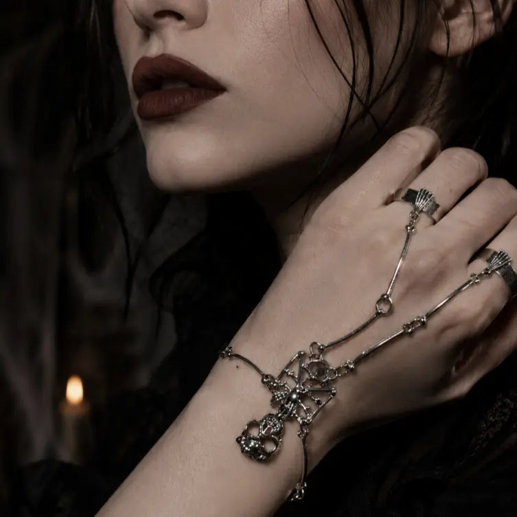 Gothic silver skeleton hand chain featuring a sculpted skull centerpiece connected to two wide-band rings, worn by a woman with dark red lipstick in a dark aesthetic setting.