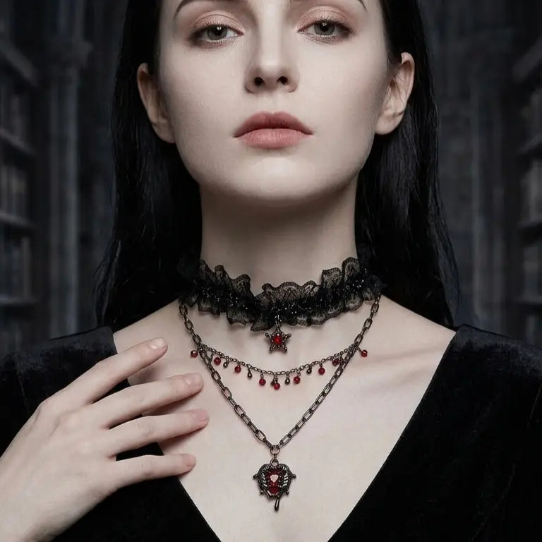 Woman with light skin tone and straight black hair wearing gothic 3-layer necklace set — black lace ruffle choker, red crystal teardrop fringe chain, and winged heart red crystal pendant chain — hand resting on chest, black V-neck top, dark gothic architectural background