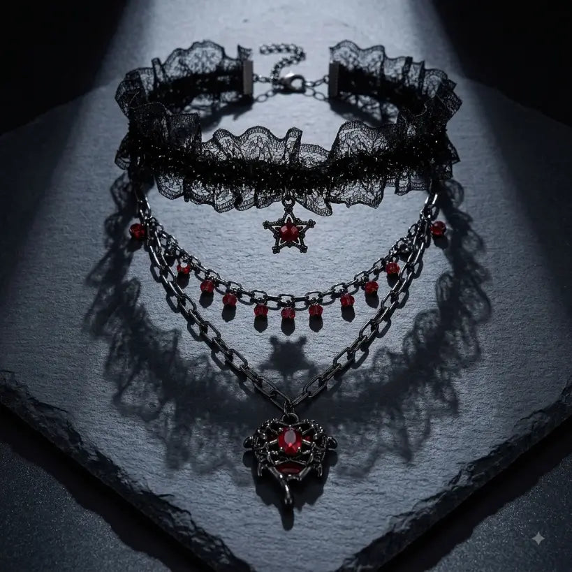 Gothic 3-layer necklace set — black lace ruffle choker with pentagram charm, red crystal teardrop fringe gunmetal chain, and filigree winged heart red crystal pendant chain — displayed on dark grey rough stone slab with dramatic single spotlight and deep shadow, black background
