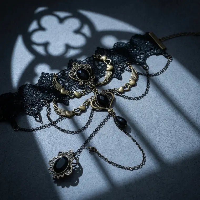 Gothic black lace angel wing onyx slave bracelet ring set with antique bronze wing charms, black faceted gemstone, draped chains with teardrop drop and connected filigree onyx ring, displayed on dark blue-grey concrete surface with dramatic gothic quatrefoil rose window and lancet arch shadow projection, strong directional spotlight, AI-generated image

