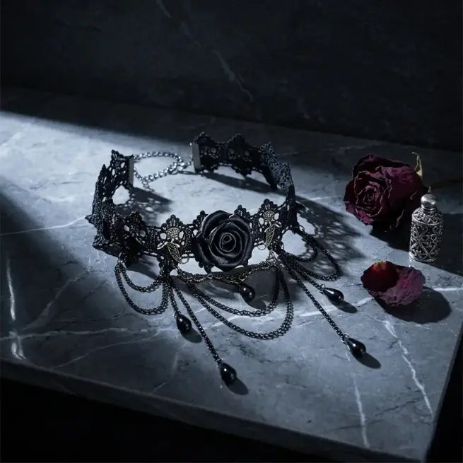 Gothic black lace rose choker necklace with draped gunmetal chains and black teardrop bead drops, displayed on dark grey marble surface with dried deep red rose, scattered petals and silver filigree perfume bottle props, dramatic single spotlight, black background, AI-generated image