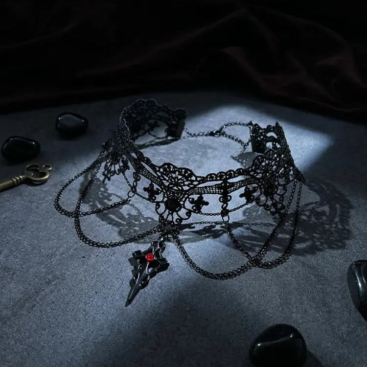 Gothic black lace fleur-de-lis filigree choker necklace with three draped gunmetal chains and black dagger spike red crystal pendant, displayed on dark grey stone slab surrounded by black obsidian pebbles and antique brass skeleton key props, dramatic single spotlight with deep shadow, black background