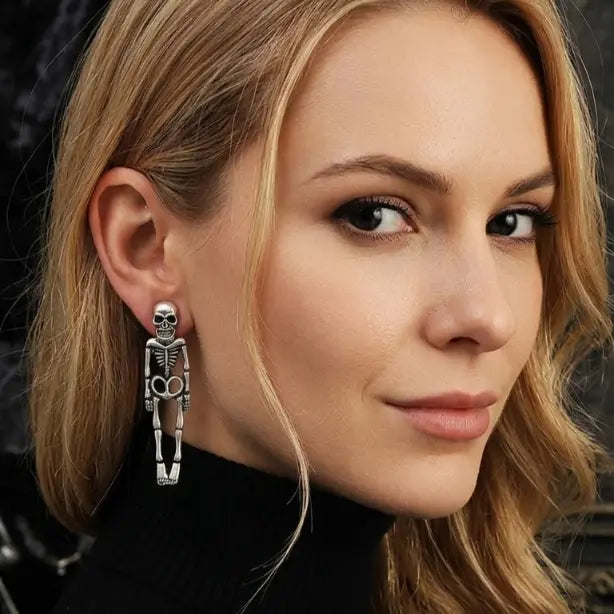 Woman with light skin tone and golden medium-length hair wearing gothic antique silver-tone full-body skeleton figure stud earring on right ear, skull head at earlobe with full bone body dangling below, black turtleneck top, dark grey marble background

