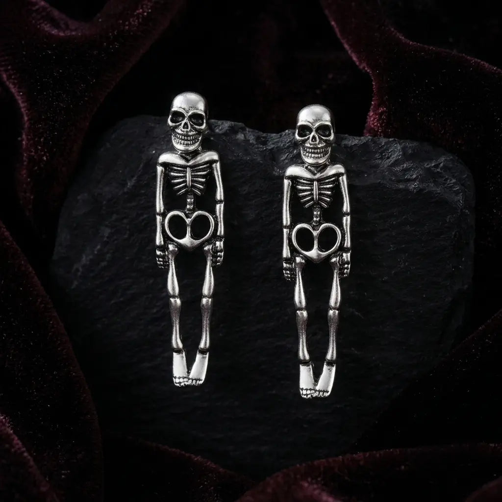 Pair of gothic antique silver-tone full-body skeleton figure stud earrings standing upright on black rough obsidian rock, deep burgundy velvet fabric background with soft natural side lighting

