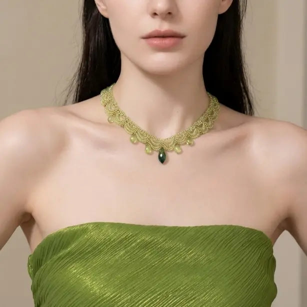 Woman with light skin tone wearing light green seed bead woven lace choker necklace with emerald green teardrop crystal pendant, dressed in olive green metallic strapless top, warm beige background

