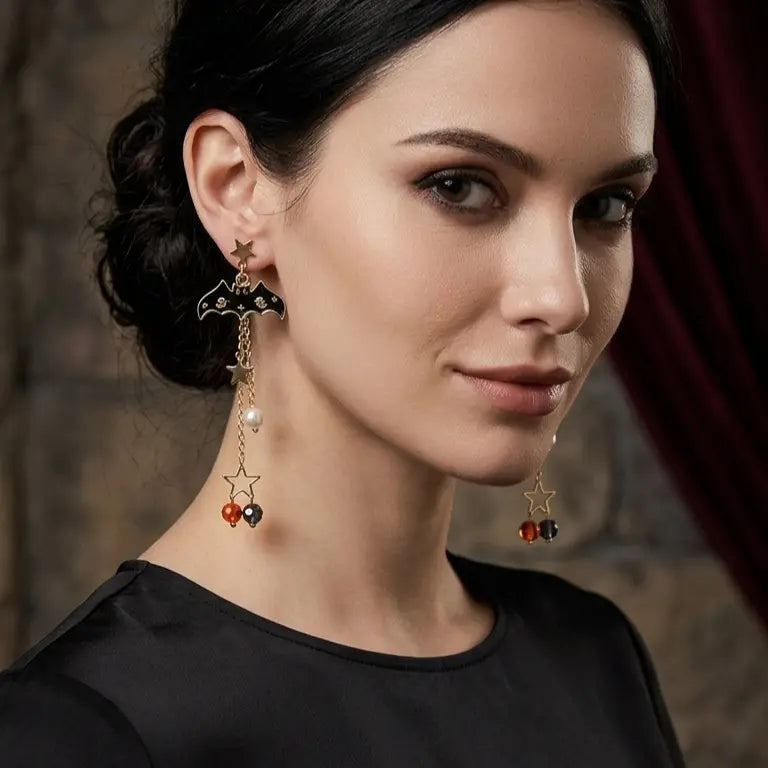 Woman with light skin tone and dark updo wearing Halloween gold-tone bat star beaded drop stud earrings — gold star stud, black enamel bat pendant, dual chain drops with pearl, open star frame and amber black crystal bead drops — black round-neck top, dark gothic stone wall with burgundy drape background

