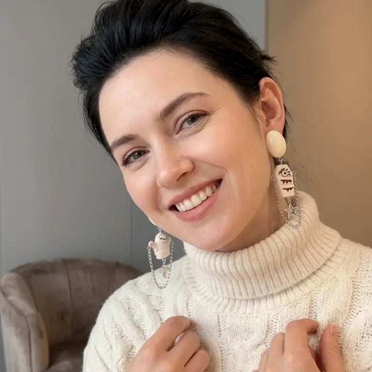 Woman with light skin tone and short black hair smiling and wearing Halloween asymmetric mummy and coffin resin drop stud earrings — mummy figure with silver chain star drop on left ear, coffin pendant with silver chain cross drop on right ear — cream chunky knit turtleneck sweater, warm grey indoor background with brown leather chair

