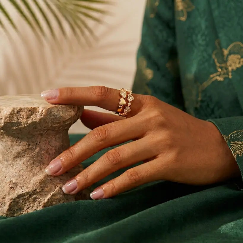 Woman wearing two stacked gold alloy heart zirconia adjustable open band rings on index finger, hand resting on travertine stone, styled with dark green embroidered satin jacket, palm leaf background