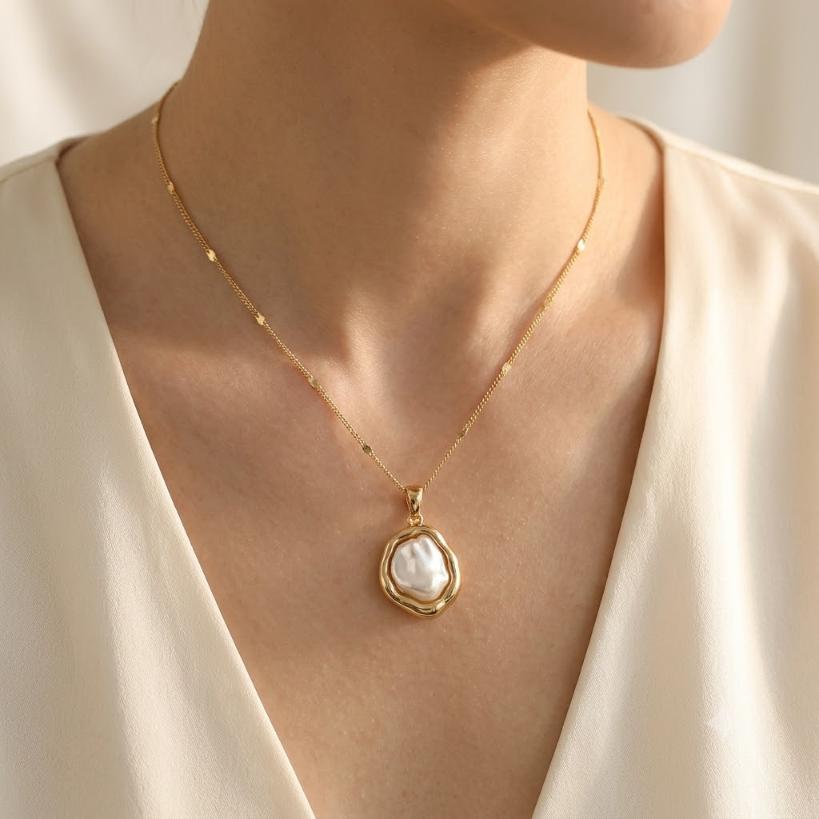 Close-up of woman's neckline wearing gold satellite chain necklace with large irregular baroque pearl pendant in wide organic gold frame, cream V-neck top, warm champagne satin background

