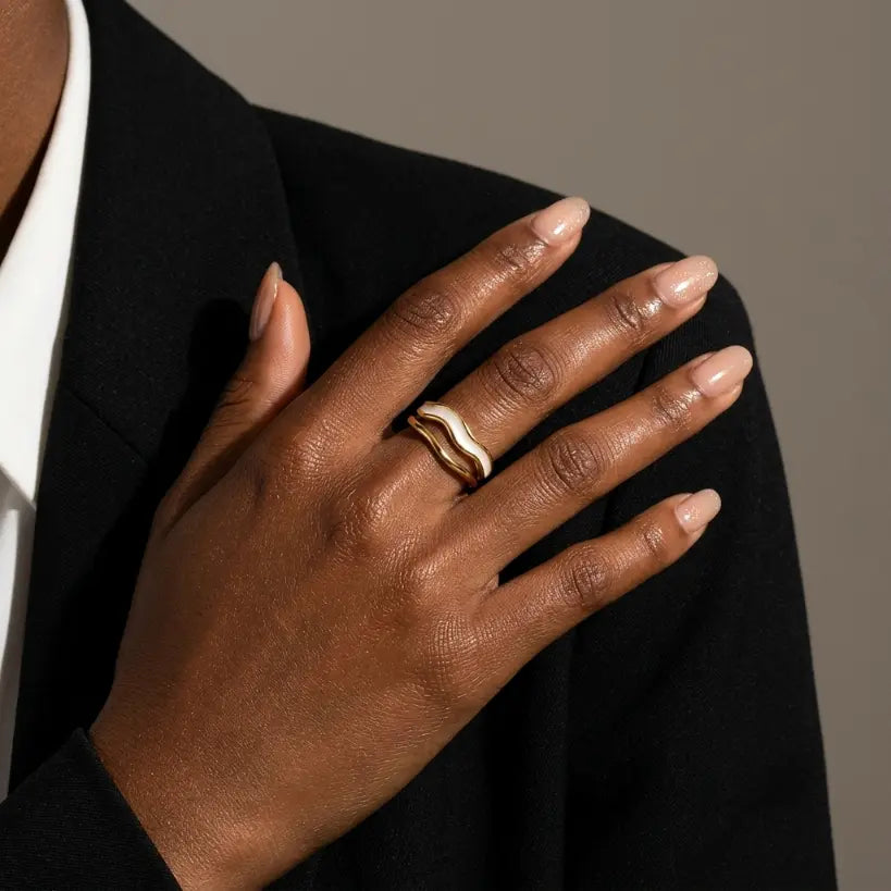Woman with deep skin tone wearing gold double-layer irregular wave open band ring on middle finger, hand resting on black blazer shoulder, styled with white shirt, warm grey background

