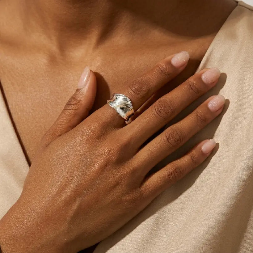 Woman with deep skin tone wearing silver copper adjustable open band irregular wide ring with hammered texture on middle finger, hand resting on chest over cream satin top in warm light

