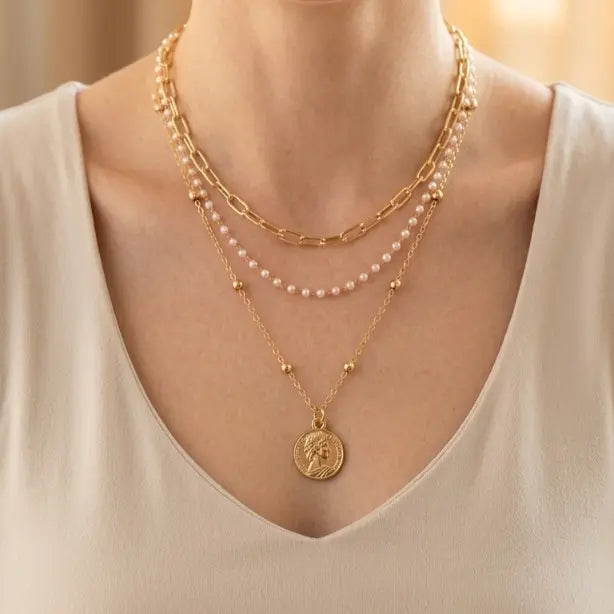Woman wearing three-layer gold necklace set — pearl and gold bead strand, paperclip chain with pearls, and satellite ball chain with large engraved portrait coin pendant — over cream V-neck top, warm golden bokeh background

