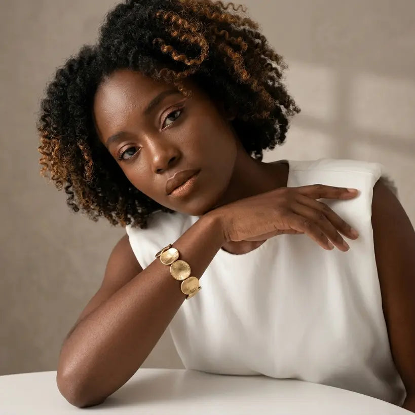 Woman with deep skin tone wearing matte gold irregular disc elastic stretch bracelet on left wrist, arm resting on shoulder, dressed in white sleeveless top, warm taupe background with diagonal window light

