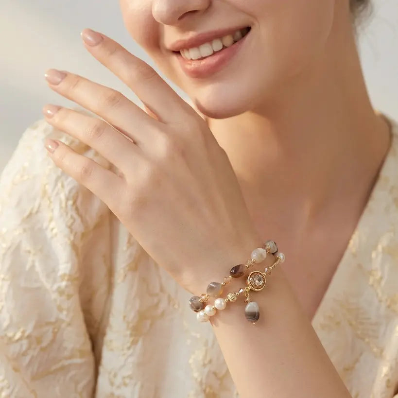 Smiling woman wearing 18K gold plated natural agate and pearl bracelet with crystal charm and agate drop pendant on left wrist, dressed in cream textured floral jacket, soft beige bokeh background

