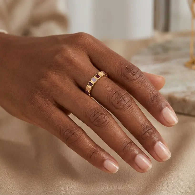 Woman with deep skin tone wearing gold alloy adjustable open band ring with alternating lavender and brown rectangular enamel tiles on middle finger, hand resting on cream satin fabric with marble surface and white jacket sleeve background

