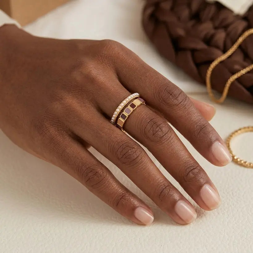 Woman with deep skin tone wearing stacked gold alloy enamel tile and freshwater pearl open band rings on middle finger, hand resting on cream surface with brown braided leather bag and gold chain in background

