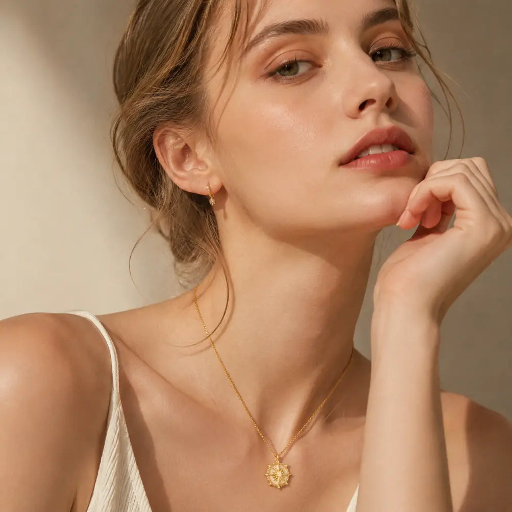 Woman with light skin tone wearing gold cable chain necklace with star compass engraved pendant, paired with gold hoop earrings, dressed in white spaghetti strap top, warm beige background with soft directional light

