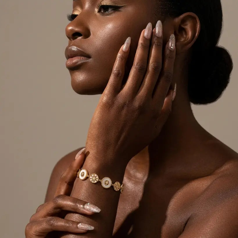 Woman with deep skin tone wearing 14K gold plated shell and zirconia flower bracelet on left wrist, hand resting against cheek, bare shoulder with glitter nail art, warm taupe background


